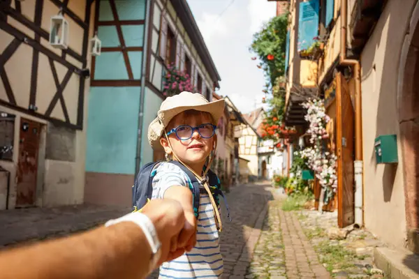 Gözlüklü ve sırt çantalı bir çocuk, Fransa 'nın Alsace bölgesinde Avrupa' da çiçeklerle süslenmiş dar bir köy caddesinde birisine rehberlik ediyor.