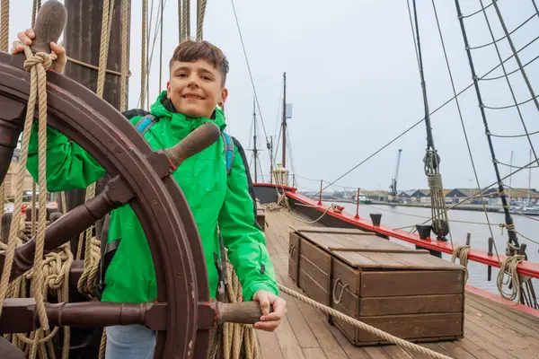 Yeşil ceketli, gülümseyen genç bir çocuk liman kıyısındaki antika bir geminin dümenini ele geçirdi.