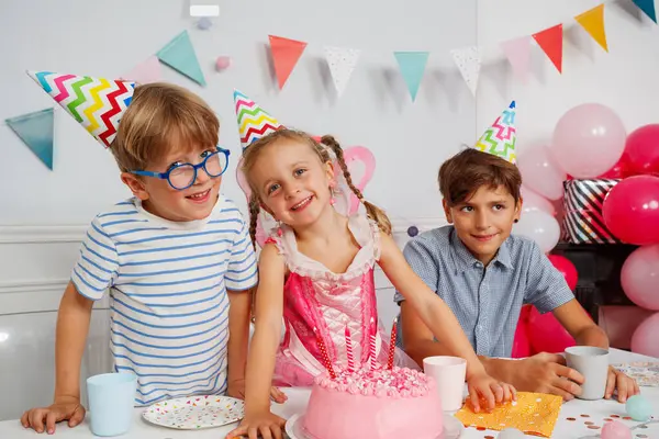 Üç çocuk renkli süslemelerle ve önünde pembe bir pastayla doğum günü kutlaması yapıyorlar. Hepsi de parlak parti şapkaları takıyor.