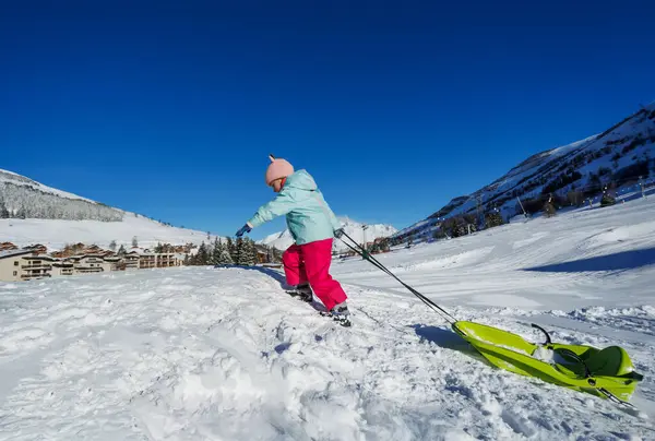 Pastel mavisi ceketli küçük bir kız tatil sırasında açık mavi gökyüzünün altında yeşil bir kızak çekiyor.