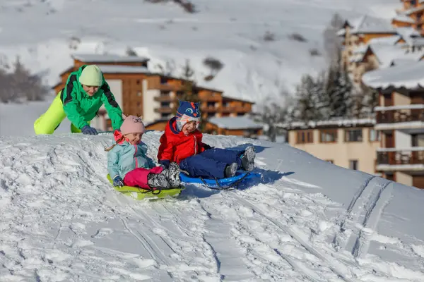 Kış mevsiminde, yeşil giyen bir çocuk diğer ikisinin kızaklara binmesine yardım eder.