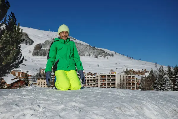 Parlak yeşil ceketli, sarı pantolonlu genç bir çocuk karla kaplı bir tepenin üzerinde gülümsüyor.