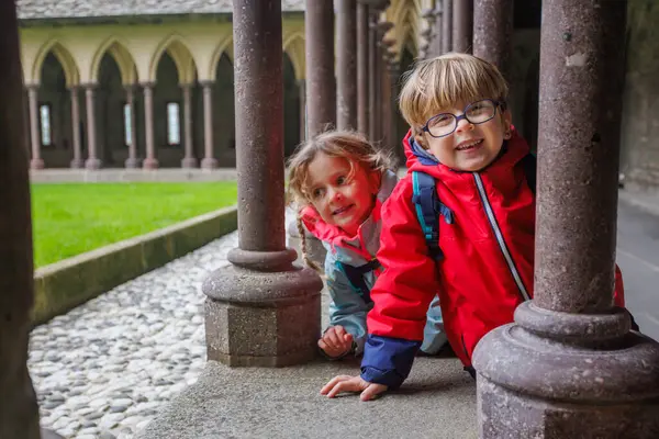 Renkli ceketler giyen iki genç çocuk, tarihi bir şato Mont Saint Michel 'de taş sütunlara yaslanırken neşeyle gülümserler.