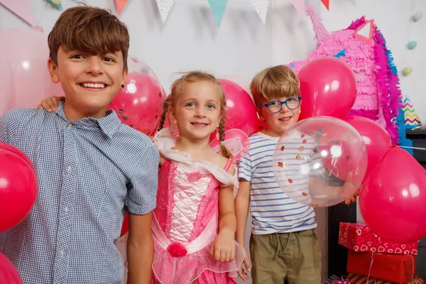 Üç çocuk canlı balonlar ve renkli süslemelerle çevrili bir kutlamada eğleniyorlar.