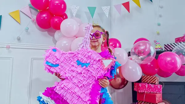 Şenlik ortamındaki küçük bir kızın elinde parlak pembe bir tek boynuzlu at pinyatası var. Renkli balonlar ve üçgen bayraklar canlı bir atmosfer yaratır.