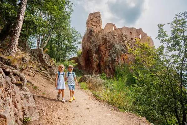 Yürüyüş yolunda duran, sırt çantalı iki çocuk geniş bir gülümseme içinde, doğa ve eski bir şatoyla çevrili.