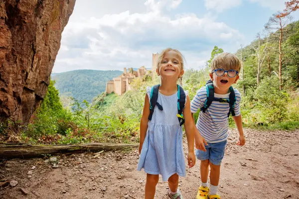 Sırt çantalı iki gülümseyen çocuk, Avrupa 'da, Fransa' nın Alsace bölgesinde arka planda antik bir şato ve yemyeşil tepeler bulunan bir patikada duruyorlar.