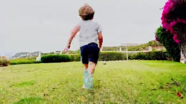 Küçük bir çocuk çimlere doğru koşuyor, bir futbol topuna doğru, etrafı palmiye ağaçlarıyla çevrili güneşli çimlerle çevrili