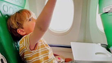 Uçakta, küçük bir çocuk mutlu bir şekilde oyuncak uçağıyla oynuyor, pencerenin yanındaki yeşil desenli koltuğun arka planında oturuyor.