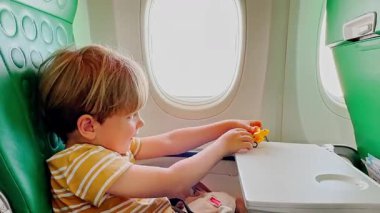 Uçakta, küçük bir çocuk mutlu bir şekilde oyuncak uçağıyla oynuyor, pencerenin yanındaki yeşil desenli koltuğun arka planında oturuyor.