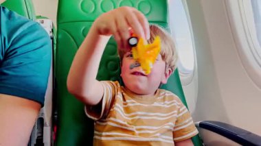 Uçakta, küçük bir çocuk mutlu bir şekilde oyuncak uçağıyla oynuyor, pencerenin yanındaki yeşil desenli koltuğun arka planında oturuyor.