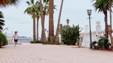 Küçük çocuk, güneşli bir günde çeşitli bitki ve palmiyelerle çevrili bir terasta koşuyor. 