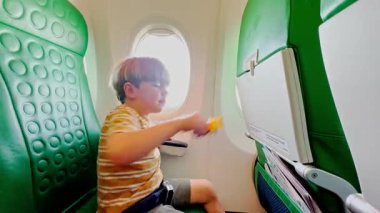 Uçakta, küçük bir çocuk mutlu bir şekilde oyuncak uçağıyla oynuyor, pencerenin yanındaki yeşil desenli koltuğun arka planında oturuyor.