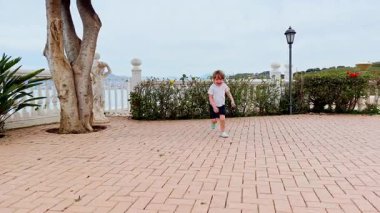 Küçük çocuk, güneşli bir günde çeşitli bitki ve palmiyelerle çevrili bir terasta koşuyor. 