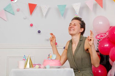 Orta yaşlı bir kadın, üzerinde pembe bir doğum günü pastası olan bir masada sevinçle oturuyor. Renkli balonlar ve kiraz kuşu arka planı süsleyin