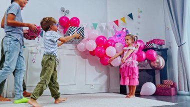 Neşeli bir kutlamada pembe elbiseli küçük bir kız iki erkekten bir hediye alır, anne, oda canlı balonlar ve çeşitli parti süsleriyle süslenir.