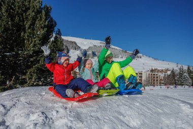 Üç çocuk bir kızağın üzerine uzanmış kollarını kaldırmış, tepenin tepesindeki bir dağ tarafından çerçevelenmiş.