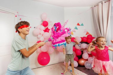 Pembe giyinmiş küçük bir kız heyecanla bakarken, gözleri bağlı bir çocuk pinataya vurmaya hazır.