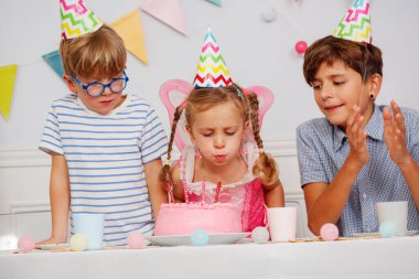 Etrafı renkli başlıklı arkadaşlarla çevrili şirin elbiseli bir kız, pembe kremalı pastadaki mumları söndürmeye hazırlanıyor.