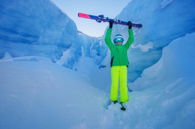 Dramatik buz kaplı uçurumlara karşı duran yeşil ceketli ve canlı pantolonlu bir kayakçı koruyucu bir kask takarak kayak pozu veriyor.