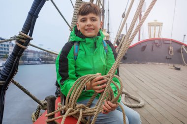 Canlı yeşil bir ceket giymiş genç bir çocuk ahşap bir gemi güvertesinde oturmuş, ipleri tutarken gülümsüyor.