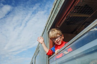 Gülümseyen bir çocuk elini hevesle uzatarak dar tren penceresinden sarkıyor.