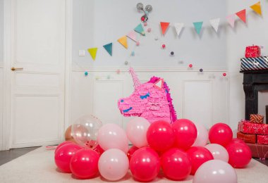 Pembe bir tek boynuzlu at pinyatası süslü bir duvarın karşısında duruyor parlak bayrak ve ışık dolu çelenklerle.