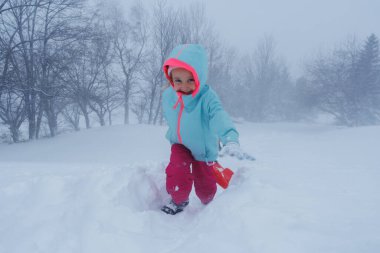 Turkuaz ceketli genç bir kız dizlerine kadar kar içinde, uzaktaki kızak ağaçlarını sürüklüyor.