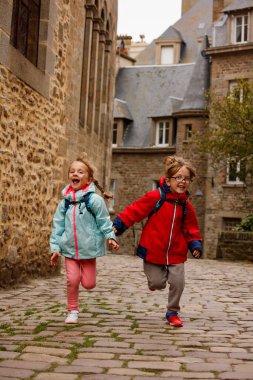 Parlak ceketler giyen iki çocuk, tarihi bir bölgede Saint-Malo, Bretagne, Fransa 'da taş binalar bulunan bir kaldırım taşından caddede neşeyle koşuyorlar.