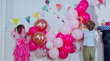 İki çocuk, erkek, kız pembe ve kırmızı balonlardan oluşan bir çelenk ile bir odayı dekore ediyor. Duvarda renkli bayraklar asılı.