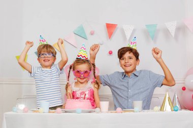 Çocuklar canlı bir kutlamada eğleniyorlar, süslü bir pasta ve renkli şapkalarla, neşeli bir atmosfer yaratıyorlar.