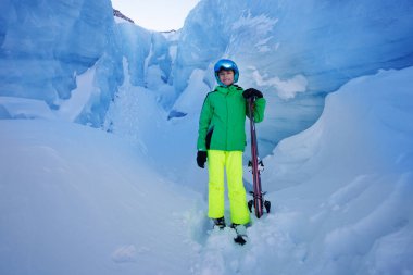 Neon yeşil bir kayak pantolonu ve parlak bir ceket giyen bir genç, yüksek mavi buz formasyonları arasında duruyor.