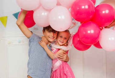 Ergen erkek kardeş doğum gününde kız kardeşine sarılıyor, elinde pembe hava balonları tutuyor.