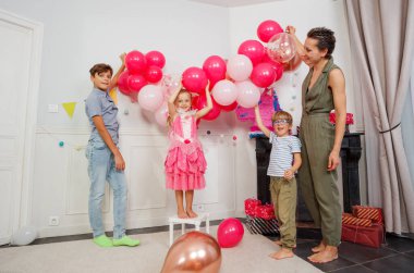 Pembe balonlarla çevrili süslü elbiseli bir kız, annesi ve erkek kardeşleri hediyeler ve kirazlarla dolu bir doğum günü atmosferi düzenlerken bir taburede duruyor.