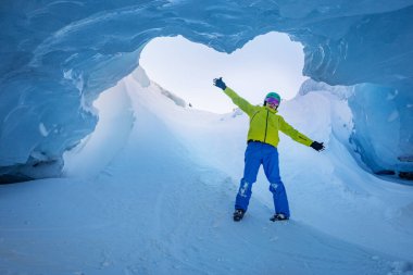 Renkli bir kayakçı, parlak mavi buz mağarasının ortasında duruyor. Tavanda gökyüzünü gösteren büyük bir delik var.