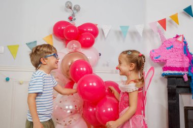 İki mutlu çocuk, gözlüklü ve çizgili tişörtlü bir çocuk, pembe peri kıyafetli şirin bir kız, doğum günü partisinde bir dizi balonla çevrili.