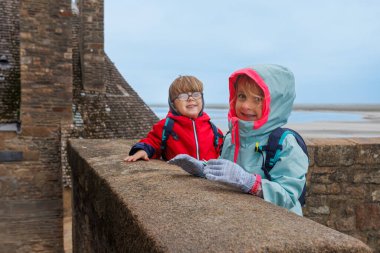 Sırt çantası takan iki çocuk sahildeki tarihi bir binanın yanındaki taş bir duvarda rüzgarlı bir günün tadını çıkarıyor.