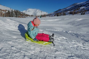 Pastel mavisi ceketli küçük bir kız arkasında dağ manzarası olan yeşil bir kızakla kayıyor.