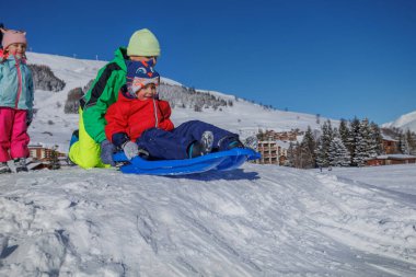 Genç kardeş, kardeşini dağdaki yamaçtan aşağıya itiyor. Kış oyunlarının tadını çıkarıyor.