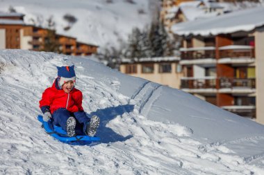 Kırmızı kar kıyafetli bir çocuk tepeden aşağı süzülüyor mavi bir kızakla, dağ evi binaları çerçeveli kar örtülü balkonlarla.