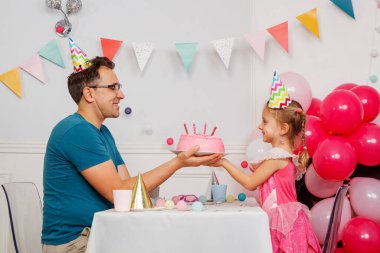 Yetişkin bir çocuk, her ikisi de parti şapkasıyla, mumları üflemek için öne doğru eğilir pembe bir doğum günü pastası üzerinde, etrafında canlı balonlar ve pankartlarla