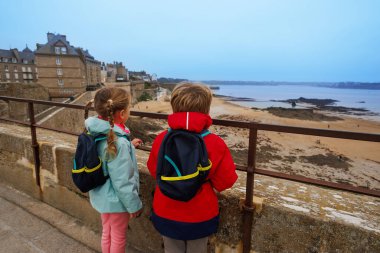 Huzurlu deniz kıyısına ve eski binalara bakan sırt çantalı iki çocuk korkulukların yanında duruyor, Saint-Malo, Bretagne, Fransa