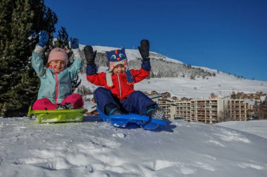 İki çocuk yeşil bir kızakla dağ evlerinin çatılarını arkalarındaki yamaçlara sürerler.