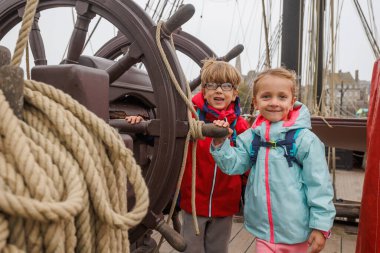 Ahşap bir güvertede, iki çocuk denizcilik iplerinin arasında, parlak dış giysiler giyip sırt çantaları taşıyorlar.