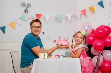 Doğum günü sahnesinde bir adam ve parti şapkalı bir kız pembe pastadaki mumları söndürmeye hazırlanıyor.