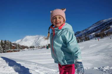 Açık mavi ceketli ve pembe şapkalı, ren geyiği kulaklı genç bir kız karlı bir manzarada oynuyor, arka planda dağlar berrak bir gökyüzünün altında