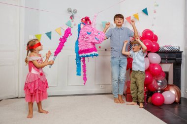 Gözleri bağlı ve prenses gibi giyinmiş genç bir kız, iki erkek, şenlikli bir odada onu neşelendirirken tek boynuzlu at pinatasına vurmaya hazırlanıyor.