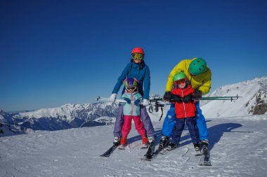 Açık mavi gökyüzüne karşı, dört kayaklı neşeli bir aile, görkemli dağlarla çevrili.