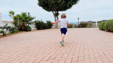 Bir çocuk, bereketli bitkiler ve ağaçlarla çerçevelenmiş, bulutlu bir gökyüzünün altındaki fayanslı terasa enerjik bir şekilde çarpar.