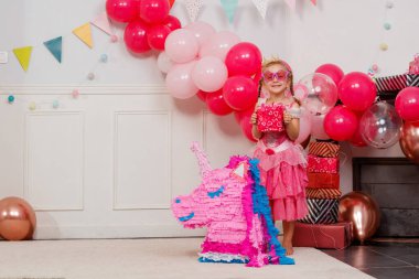 Şenlikli bir mekanda, prenses elbisesi giymiş genç bir kız, parlak bir tek boynuzlu atın yanında poz veriyor.